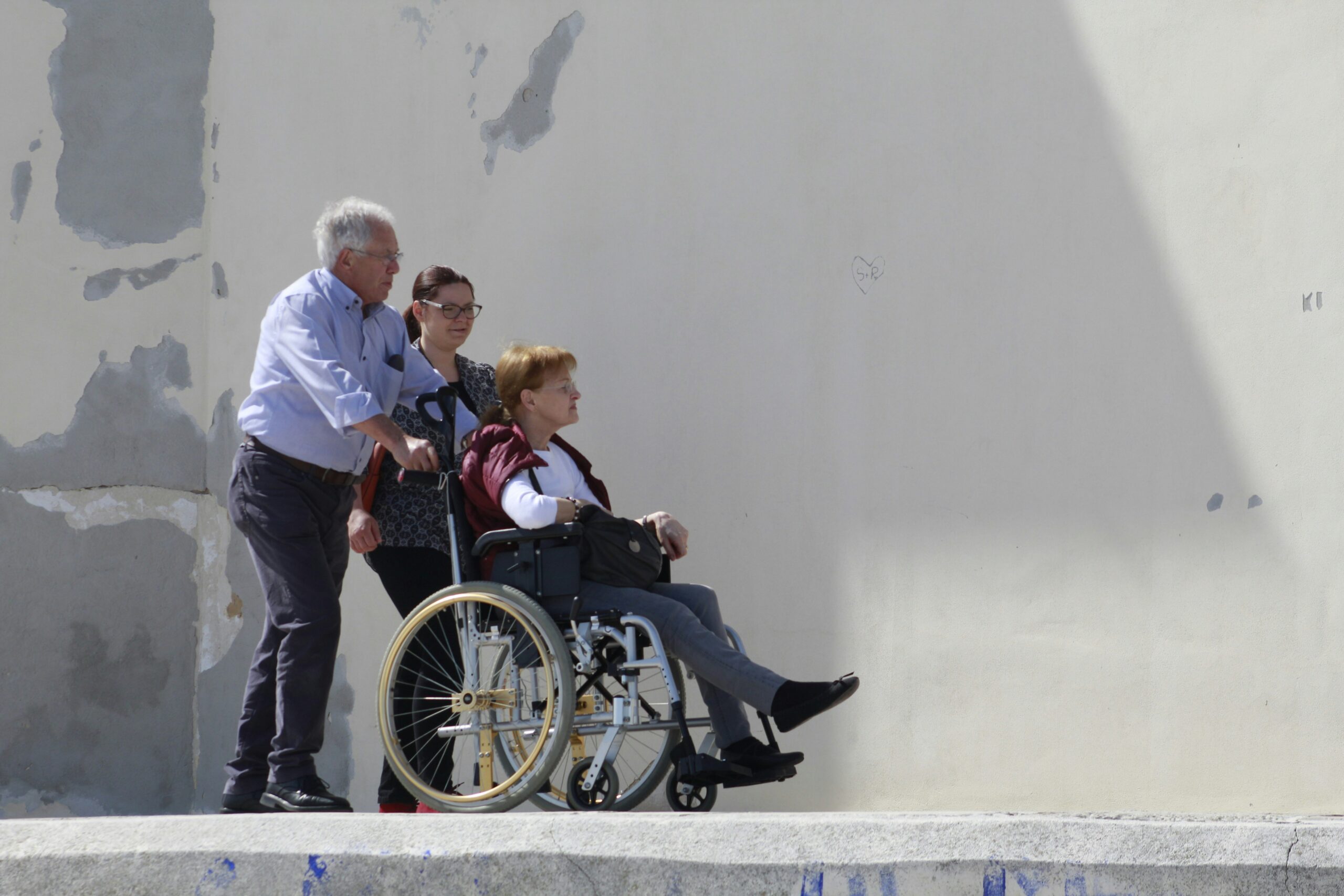 vrouw in rolstoel geduwd door man en vrouw voor grijze muur